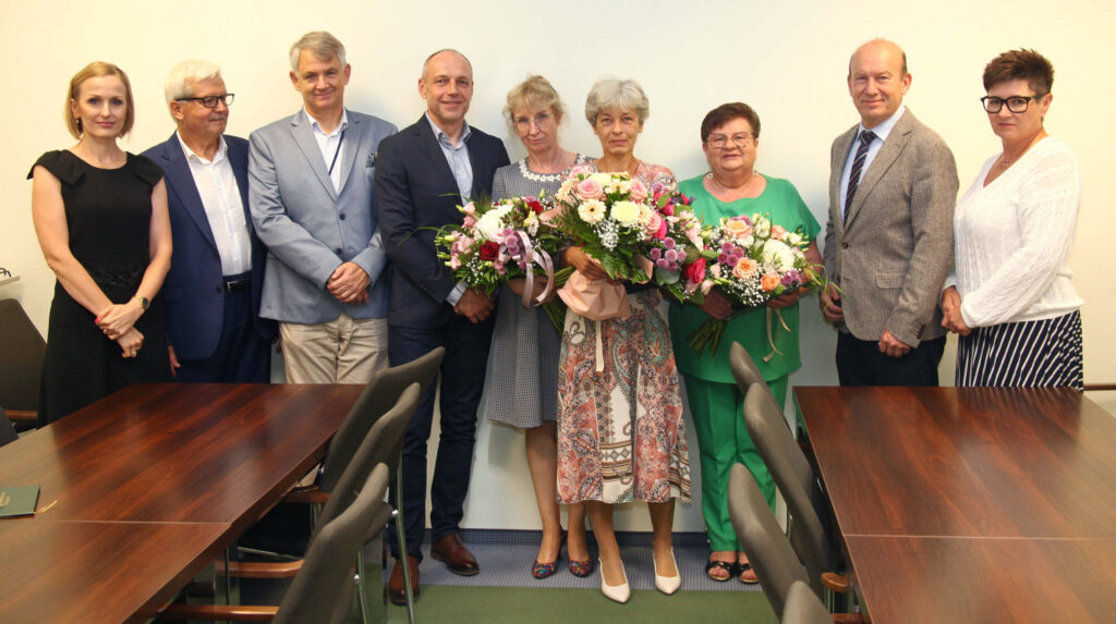 grupa uśmiechniętych osób w środku trzy panie trzymają bukiety kwiatów